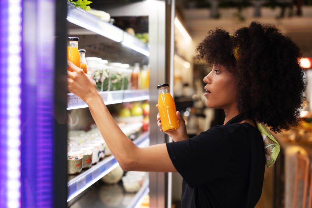 Side view of black female buyer standing at counter in supermarket and picking orange juice in bottle