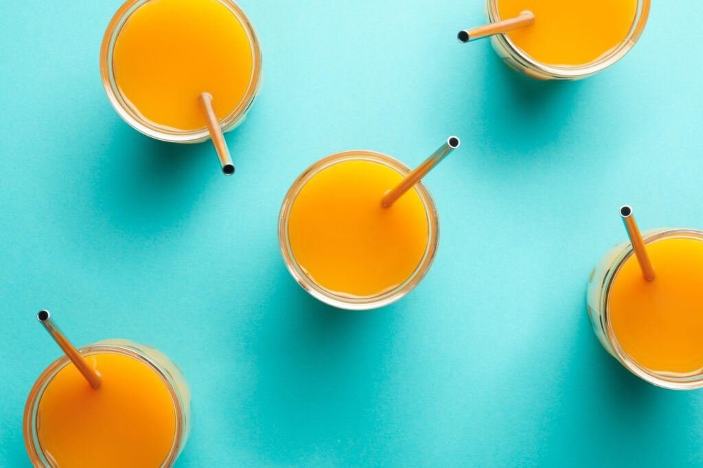 Top view of arranged glasses with orange beverage and reusable steel straws on blue background