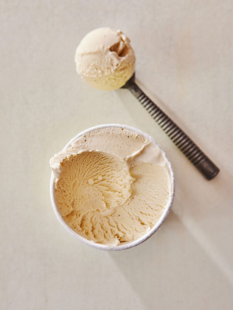 Open paper carton of vanilla ice cream. Laying next to ice cream tub, a metal ice cream scoop with perfect round ball of ice cream. White background.