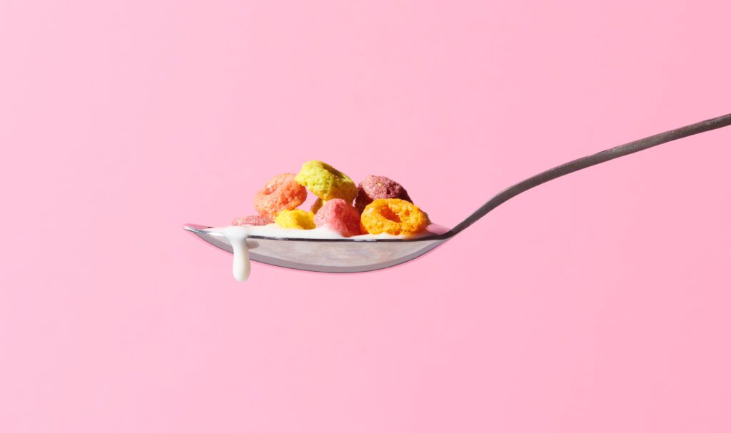 Multicoloured fruit cereal with dripping milk on spoon against pink background