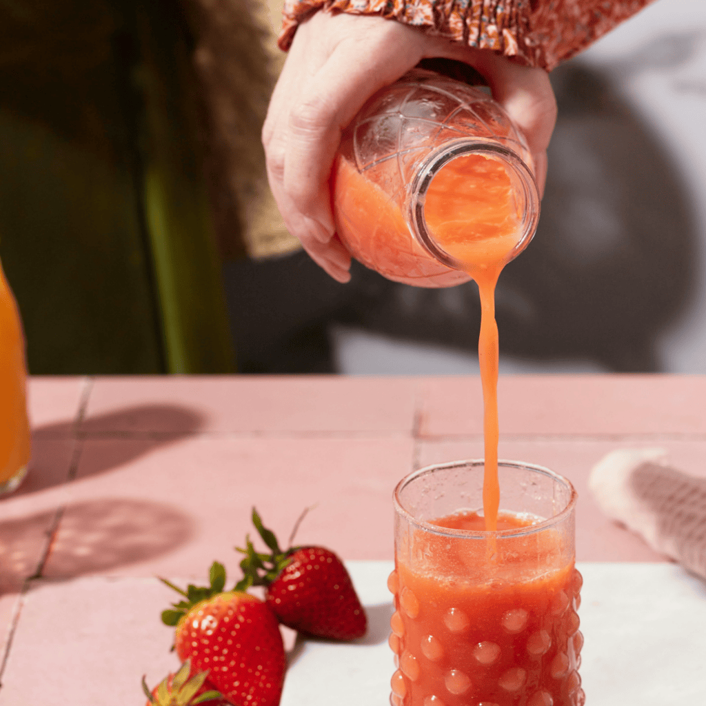 A strawberry-pink beverage being poured out of a glass bottle into a glass cup sitting on a pink counter.